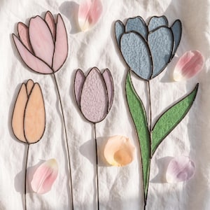 Op de afbeelding: Drie glas-in-lood tulp ornamenten met metalen soldeerlijnen. Eén tulp is lichtroze, één is perzikkleurig en één is lichtpaars. Een grotere blauwe glas-in-lood tulp met een groene steel en bladeren is ook aanwezig, omringd door losse bloemblaadjes.