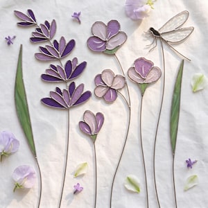 May include: A collection of stained glass art pieces featuring a tall purple lavender stalk, three smaller purple and light pink flowers on thin stems, and a dragonfly with clear wings, all accented by two long green leaves and scattered petals.