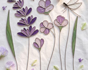 May include: A collection of stained glass art pieces featuring a tall purple lavender stalk, three smaller purple and light pink flowers on thin stems, and a dragonfly with clear wings, all accented by two long green leaves and scattered petals.