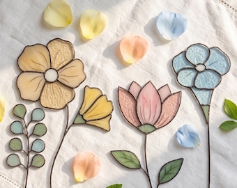 May include: A collection of stained glass flowers and a leafy stem featuring a large yellow blossom, a pink tulip-like flower, and a light blue flower, all with dark metal soldering lines, accompanied by scattered loose flower petals.