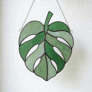 May include: A stained glass leaf in shades of green, with a dark border and a chain for hanging. The leaf design features cut-out sections, creating an open, airy look. The glass has varying textures and tones, adding depth to the piece.