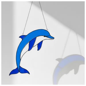 May include: A blue and white stained glass dolphin, hanging from a silver chain. The dolphin's design is streamlined, with a black eye. The image is set against a white backdrop, with the dolphin's shadow visible.