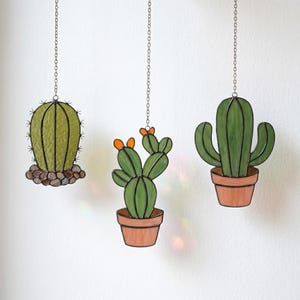 Op de afbeelding: Drie hangende cactusdecoraties van glas-in-lood aan zilveren kettingen. De linker cactus is rond met bruine glazen steentjes aan de basis. De middelste cactus is gesegmenteerd met oranje bloemen in een terracotta pot. De rechter is hoog.