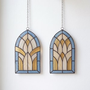 Könnte beinhalten: Zwei bogenförmige Buntglasfenster mit einem geometrischen Muster aus hellblauen, cremefarbenen und goldenen Glassegmenten, die durch dunkle Metallstege verbunden sind. Jedes Element hängt an einer silberfarbenen Metallkette.
