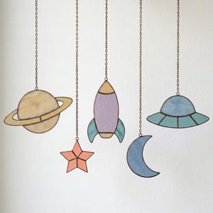May include: Five stained glass space-themed ornaments hanging from chains. The ornaments include a planet with rings, a star, a rocket, a crescent moon, and a UFO. Each ornament is made of colorful glass and outlined with a dark metal.