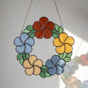 Puede incluir: Una corona floral de vidrieras con cinco flores en tonos azul, amarillo y naranja, rodeadas de hojas verdes. La corona está suspendida por una cadena plateada, un elemento decorativo.