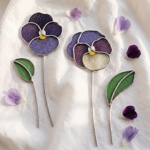 Puede incluir: Dos flores de pensamiento de vitral con tallos de metal delgados y hojas verdes. Una flor presenta pétalos de color púrpura intenso con un centro amarillo, mientras que la otra combina pétalos púrpuras y crema con un centro amarillo y detalles lineales.