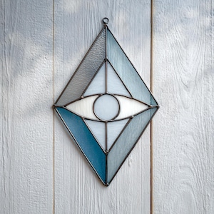May include: A stained glass wall hanging in a diamond shape, featuring an eye design. The artwork uses shades of blue, white, and gray glass, with a dark metal frame. The eye's pupil is a white circle.