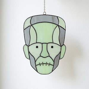May include: A stained glass suncatcher shaped like a stylized head with pale green and grey glass panels held together by black metal solder lines. The design features two small black circular eyes and a horizontal line across the mouth with vertical stitches.