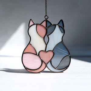 Könnte beinhalten: Glasfenster-Suncatcher mit zwei Katzen in Rosa, Weiß und Grau, verbunden durch ein rosa Herz. Die Umrisse sind schwarz, und es hängt an einer Kette, ideal für Fenster- oder Wanddekoration.