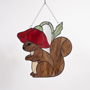 Può includere: Un acchiappasole in vetro colorato raffigurante uno scoiattolo marrone con il ventre color crema e una grande coda folta. Lo scoiattolo è posizionato sotto un papavero rosso brillante con stelo e foglia verde chiaro, appeso a una catenella argentata.