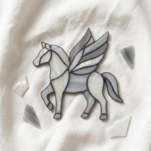 Op de afbeelding: Een glas-in-lood pegasus ornament gemaakt van iriserende witte en doorschijnende grijze glasstukken verbonden door donkere metalen soldeerlijnen. De gevleugelde paardfiguur is omringd door vier kleine, losse scherven van bijpassend glas.