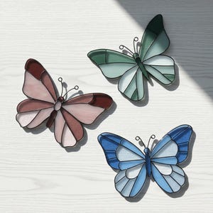 May include: Three stained glass butterfly decorations in pink, green, and blue. Each butterfly has a black outline and delicate antenna details. The butterflies are arranged on a white wooden surface.