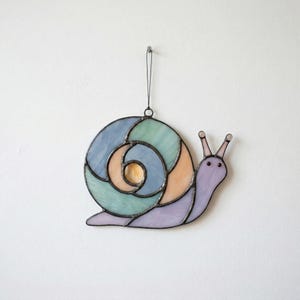 May include: A colourful stained glass snail decoration. The shell is a spiral of blue, green, and peach panels. The snail's body is lavender. It has two small antennae and a black hanging string.