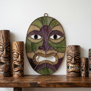 May include: A display of Tiki-themed decorations. A stained glass mask in green, purple, and tan hangs above carved wooden Tiki statues. The statues are of varying sizes and feature detailed facial expressions.