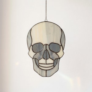 May include: A stained glass skull decoration, hanging from a chain. The skull is composed of various shades of white, gray, and clear glass, with dark lines defining the features. The design is symmetrical and detailed.