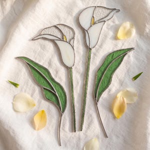 Pode incluir: Duas flores de copo-de-leite feitas de vitral com pétalas brancas, centros amarelos e folhas verdes. Cada flor é construída com linhas de metal soldadas que mantêm as peças de vidro unidas, criando uma peça de arte botânica decorativa.