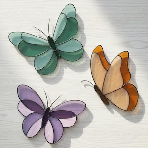 Può includere: Tre decorazioni a farfalla in vetro colorato nei toni del verde, arancione e viola. Ogni farfalla ha un contorno nero e delicate antenne. Le farfalle sono disposte su una superficie di legno chiaro.