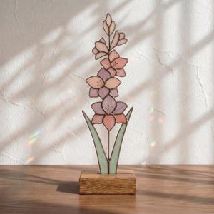 May include: A stained glass gladiolus flower sculpture featuring multiple petals in shades of soft pink, mauve, and lavender. The glass flower is supported by a thin metal stem and two pale green leaves, all mounted on a rectangular wooden base.