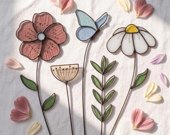 May include: A collection of stained glass garden stakes featuring a large dusty rose flower, a light blue butterfly, a small fan-shaped bloom, a green leafy stem, and a white daisy with a yellow center, all surrounded by scattered flower petals.