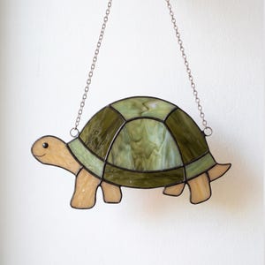 May include: A stained glass turtle suncatcher featuring a shell made of variegated olive and sage green glass panels. The head, legs, and tail are crafted from light beige glass. The pieces are joined by dark metal solder and hang from a silver chain.