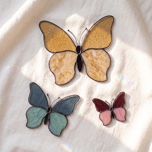 May include: Three stained glass butterfly ornaments with dark metal soldering. The largest butterfly has yellow and cream wings, the medium one has blue and light green wings, and the smallest one features deep red and light pink wing sections.