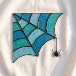 May include: A stained glass spider web panel featuring various shades of blue and teal glass segments joined by dark metal solder lines. A small, dark metal spider figure hangs from the bottom right corner of the web by a thin metal wire.