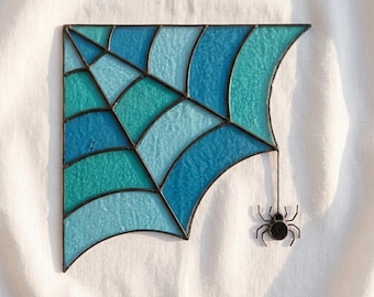 May include: A stained glass spider web panel featuring various shades of blue and teal glass segments joined by dark metal solder lines. A small, dark metal spider figure hangs from the bottom right corner of the web by a thin metal wire.