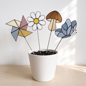 May include: Four stained glass garden stakes featuring a multi-colored pinwheel, a white daisy with a yellow center, a brown mushroom, and a light blue butterfly, all inserted into a white ceramic pot filled with dark potting soil.