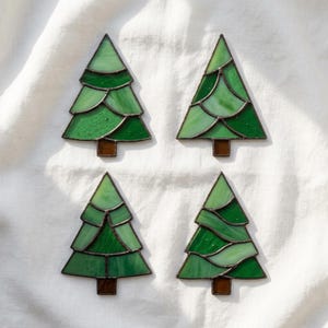 May include: Four stained glass ornaments shaped like evergreen trees. Each tree features layered segments of light and dark green glass joined by dark metal solder lines, with a small rectangular brown glass base at the bottom of each piece.