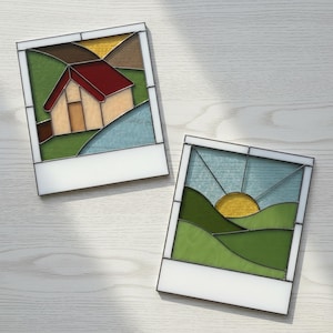 May include: Two stained glass art pieces in the shape of polaroid photos. One depicts a house with a red roof and green hills. The other shows a sunset over green hills. Both have white borders.