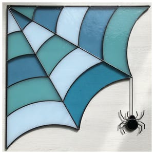 Puede incluir: Telaraña de vidriera en tonos azules y blancos, con una araña negra colgando debajo. El diseño de la telaraña presenta segmentos curvos y un borde negro, ideal para Halloween.