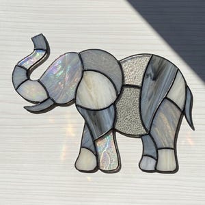 以下が含まれることがあります： グレー、白、虹色のガラスを使ったステンドグラスのゾウの彫刻。ゾウは鼻を上げており、幾何学的な形で構成されています。ガラスは質感があり、黒い輪郭があります。