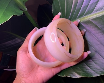 May include: Two white bangle bracelets with a subtle marbled pattern. The bracelets are stacked on top of each other.