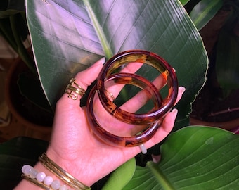May include: Two brown and amber plastic bangles stacked on top of each other. The bangles are round and have a glossy finish.