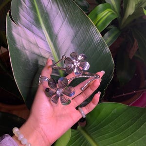 May include: Silver floral bracelet with two flower charms. The bracelet is adjustable and has a delicate design.