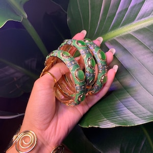 May include: Three gold bangles with green enamel and round green stones. The bangles are stacked on top of each other.