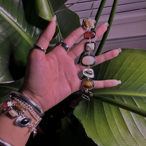 May include: A silver bracelet with a series of oval and rectangular stones in shades of brown, orange, and white. The bracelet is held up against a backdrop of green leaves. Several other silver bracelets and rings are also visible.