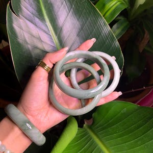 May include: A collection of jade bracelets in varying shades of green. The bracelets are round and smooth, with a polished finish. One bracelet is worn on the wrist, while others are held in the hand. The background features lush green foliage.