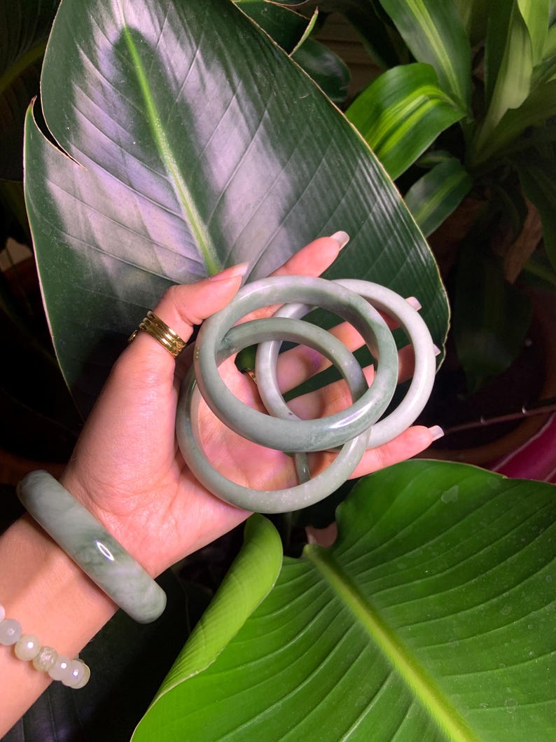 May include: A collection of jade bracelets in varying shades of green. The bracelets are round and smooth, with a polished finish. One bracelet is worn on the wrist, while others are held in the hand. The background features lush green foliage.
