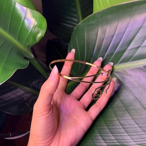 Peut inclure: Un bracelet en m&eacute;tal dor&eacute; avec un motif en spirale. Le bracelet est tenu dans une main sur un fond de feuilles vertes.