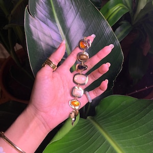 May include: A gold bracelet with a series of colorful, oval-shaped stones. The stones are set in gold and are arranged in a row. The bracelet is on a person's wrist.