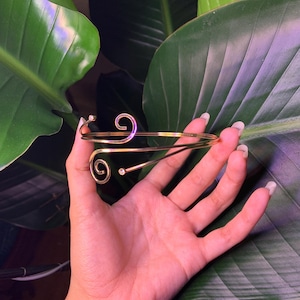 Peut inclure: Un bracelet en m&eacute;tal dor&eacute; avec un design d&eacute;licat et tourbillonnant. Le bracelet a deux petites pierres claires.