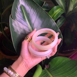 May include: Two white bangle bracelets with a gold stripe, stacked on top of each other. The bracelets are made of a smooth, polished material.