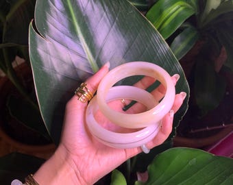 May include: Two white bangle bracelets with a gold stripe, stacked on top of each other. The bracelets are made of a smooth, polished material.