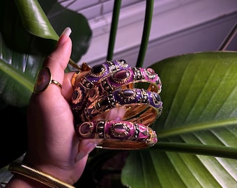 Op de afbeelding: Een verzameling goudkleurige armbanden en ringen. De armbanden hebben ingewikkelde ontwerpen met paarse en roze accenten. Een gouden ring met een grote, ovale steen is ook zichtbaar. De sieraden worden gepresenteerd tegen een achtergrond van groene bladeren.