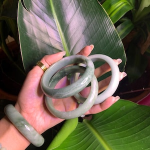 May include: A hand displays several jade bracelets in varying shades of green. The bracelets are smooth and polished, with a natural, marbled appearance. The background features large, green leaves, creating a natural and elegant aesthetic.