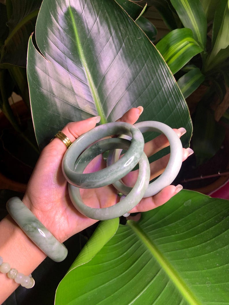 May include: A hand displays several jade bracelets in varying shades of green. The bracelets are smooth and polished, with a natural, marbled appearance. The background features large, green leaves, creating a natural and elegant aesthetic.