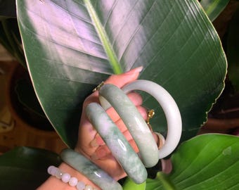 May include: A collection of four smooth, rounded jade bangles in varying shades of pale green, mottled green, and solid light gray, accompanied by a single beaded bracelet featuring small, round, translucent white beads on a flexible cord.