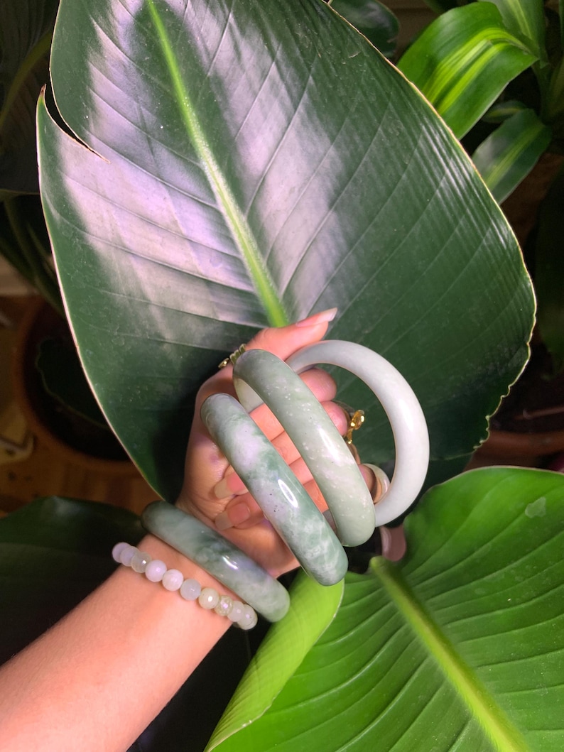 May include: A collection of jade bracelets in varying shades of green and white. The bracelets are displayed on a wrist, with a beaded bracelet also visible. The background features large green leaves.